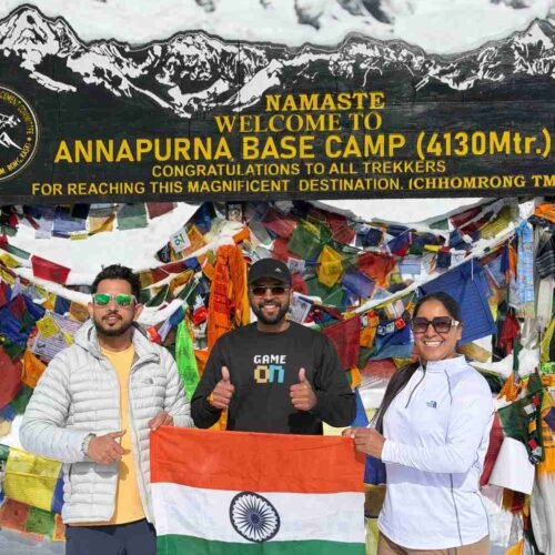 Three trekkers holding the Indian flag at Annapurna Base Camp (4,130 m) in Nepal, standing in front of colorful prayer flags and a snow-covered mountain backdrop.