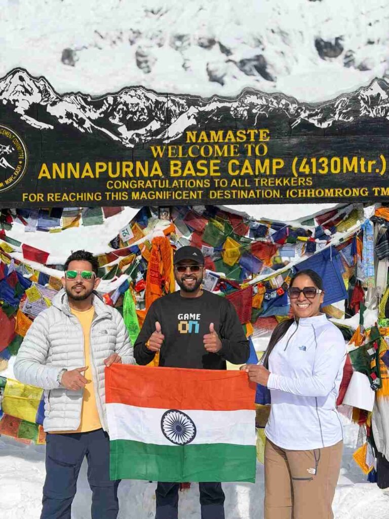 Three trekkers holding the Indian flag at Annapurna Base Camp (4,130 m) in Nepal, standing in front of colorful prayer flags and a snow-covered mountain backdrop.