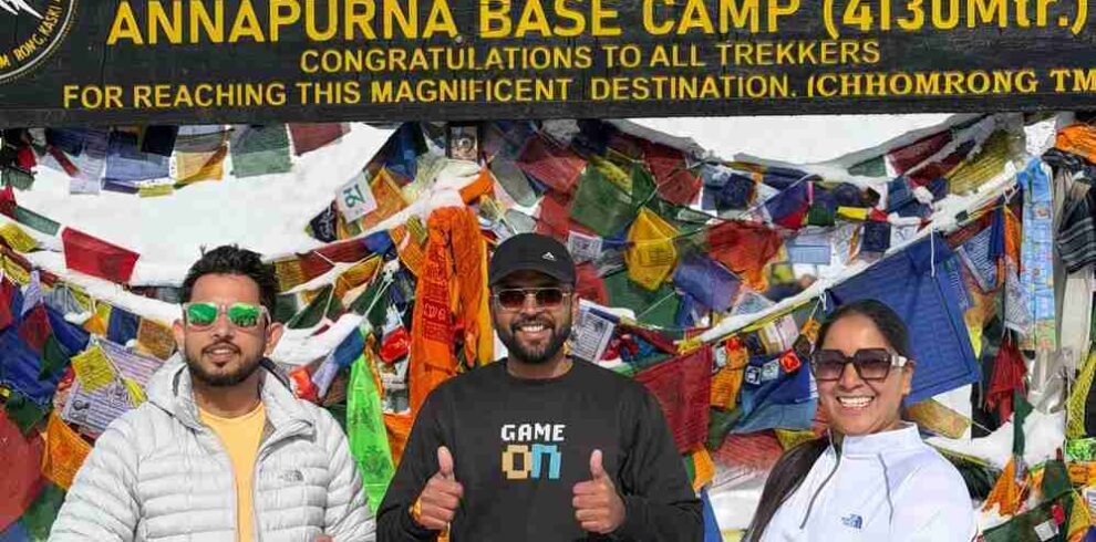 Three trekkers holding the Indian flag at Annapurna Base Camp (4,130 m) in Nepal, standing in front of colorful prayer flags and a snow-covered mountain backdrop.