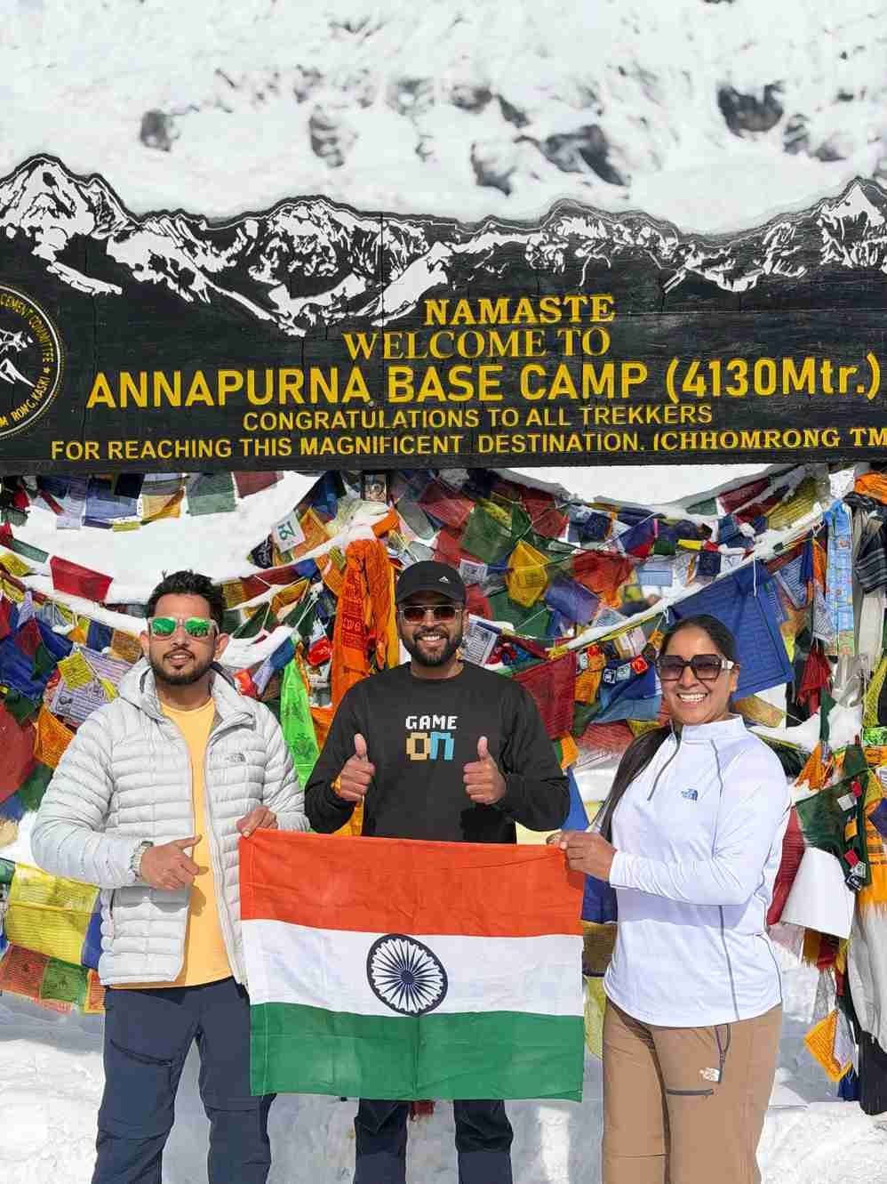 Three trekkers holding the Indian flag at Annapurna Base Camp (4,130 m) in Nepal, standing in front of colorful prayer flags and a snow-covered mountain backdrop.