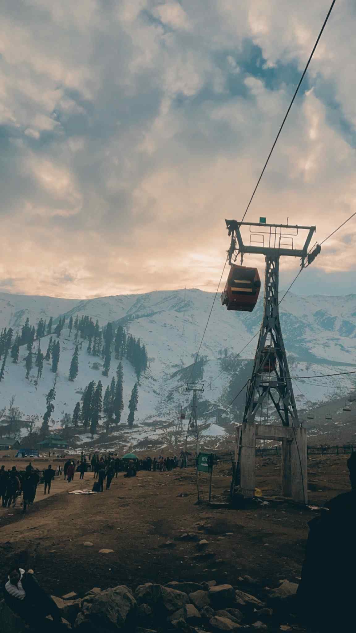A gondola cable car gliding above a mountain valley with snow-covered slopes under a dramatic evening sky. 