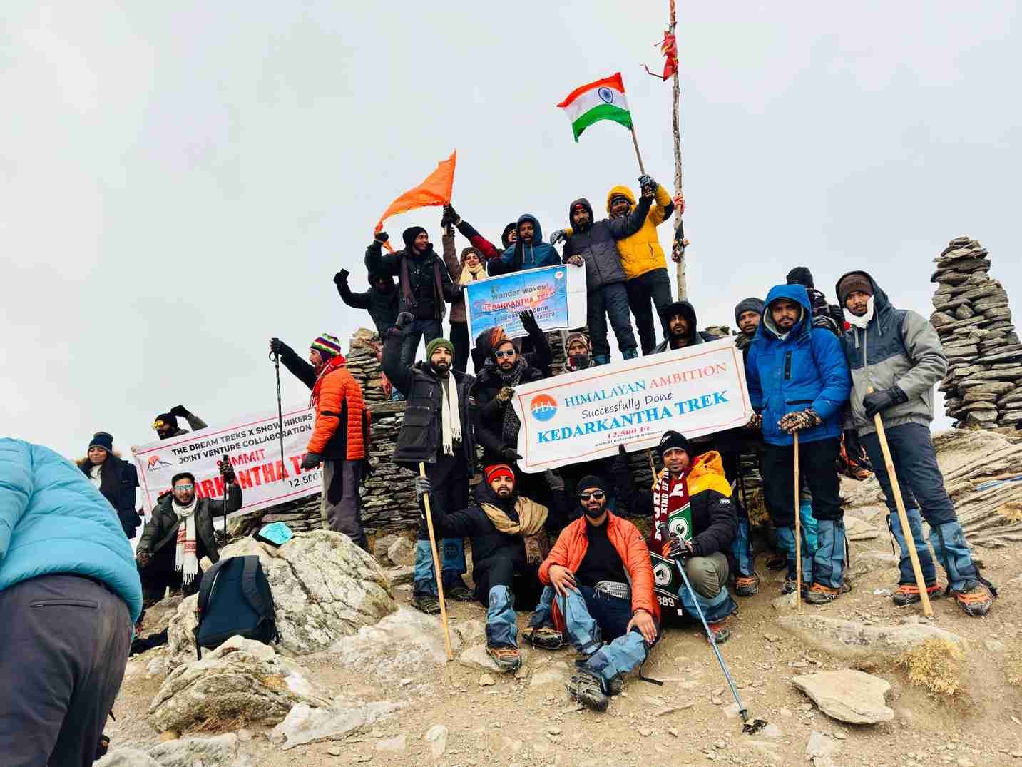 A group of trekkers celebrating at the Kedarkantha summit, holding flags and banners against a rugged mountain backdrop. 