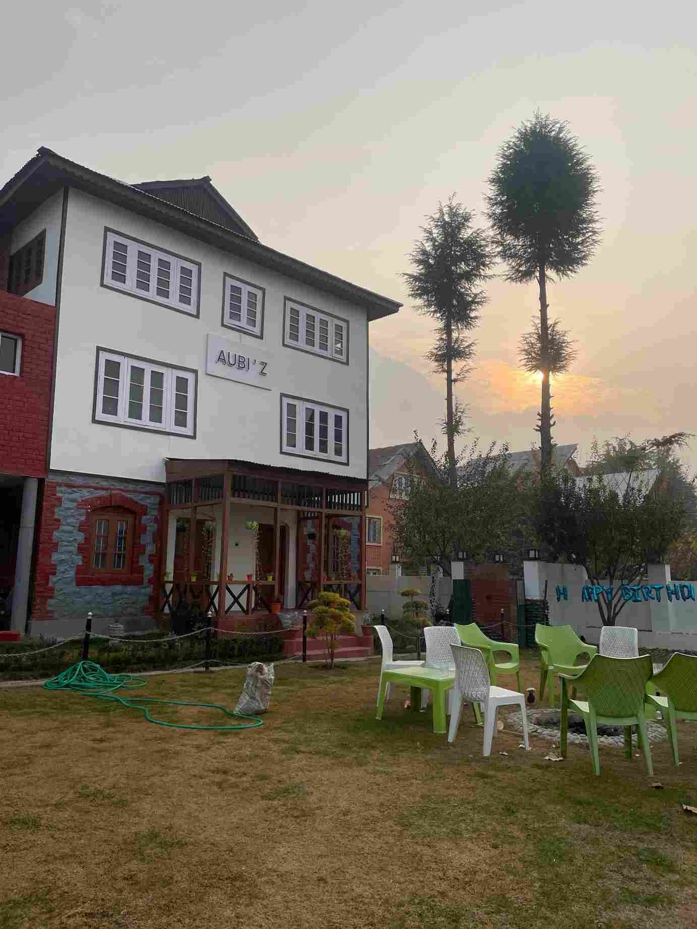 A cozy mountain guesthouse with a garden seating area, illuminated by the warm glow of a sunset behind tall trees. 