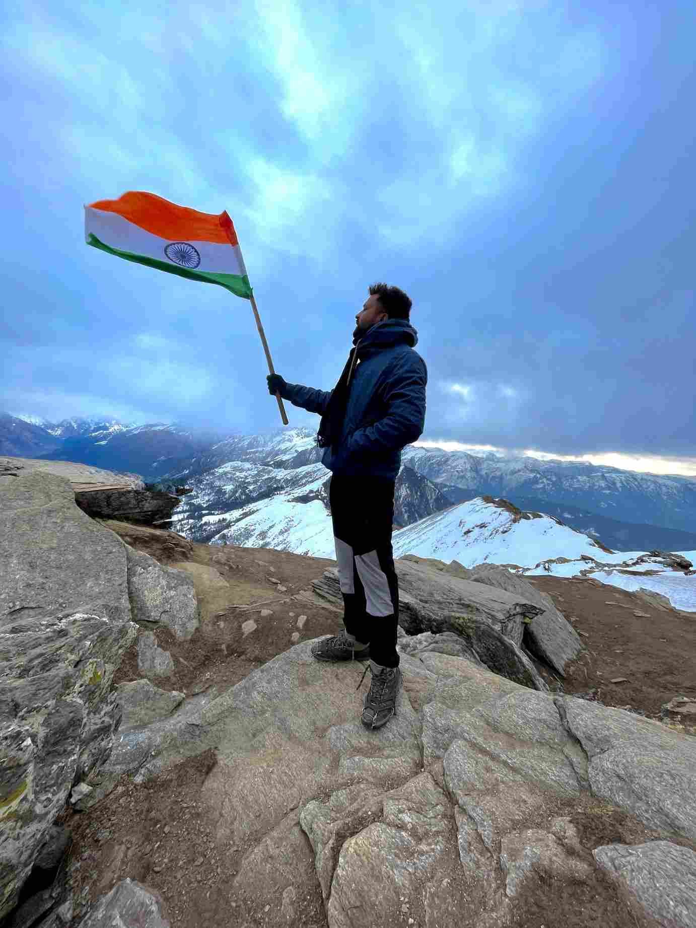 A traveler standing on a rocky mountain peak holding the Indian national flag, with snow-covered Himalayan ranges and dramatic clouds in the background. 