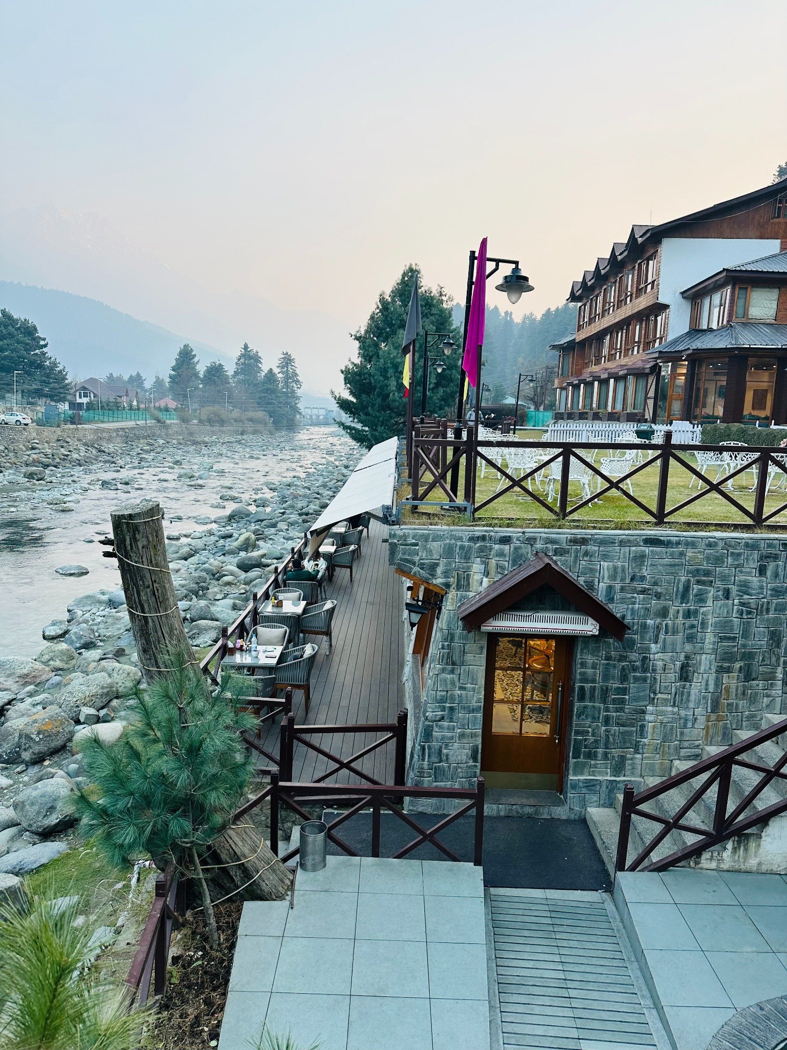 A scenic riverside resort with outdoor seating, surrounded by pine trees and misty mountain views. 