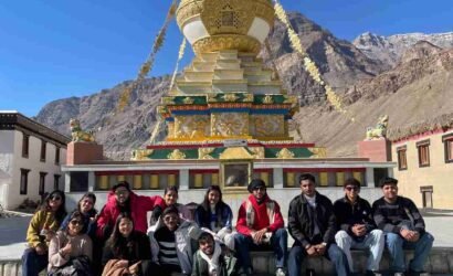 A group of friends posing in front of a golden Buddhist stupa in a Himalayan mountain village under a clear blue sky.