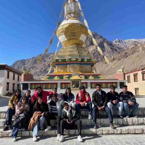 A group of friends posing in front of a golden Buddhist stupa in a Himalayan mountain village under a clear blue sky.