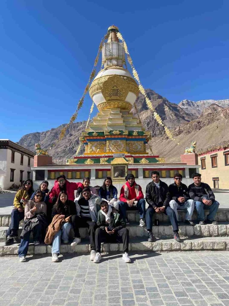 A group of friends posing in front of a golden Buddhist stupa in a Himalayan mountain village under a clear blue sky.