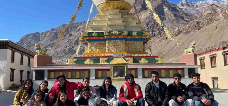 A group of friends posing in front of a golden Buddhist stupa in a Himalayan mountain village under a clear blue sky.