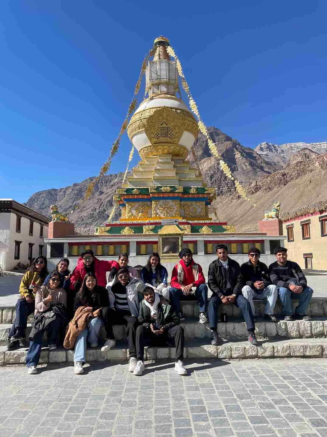 A group of friends posing in front of a golden Buddhist stupa in a Himalayan mountain village under a clear blue sky.