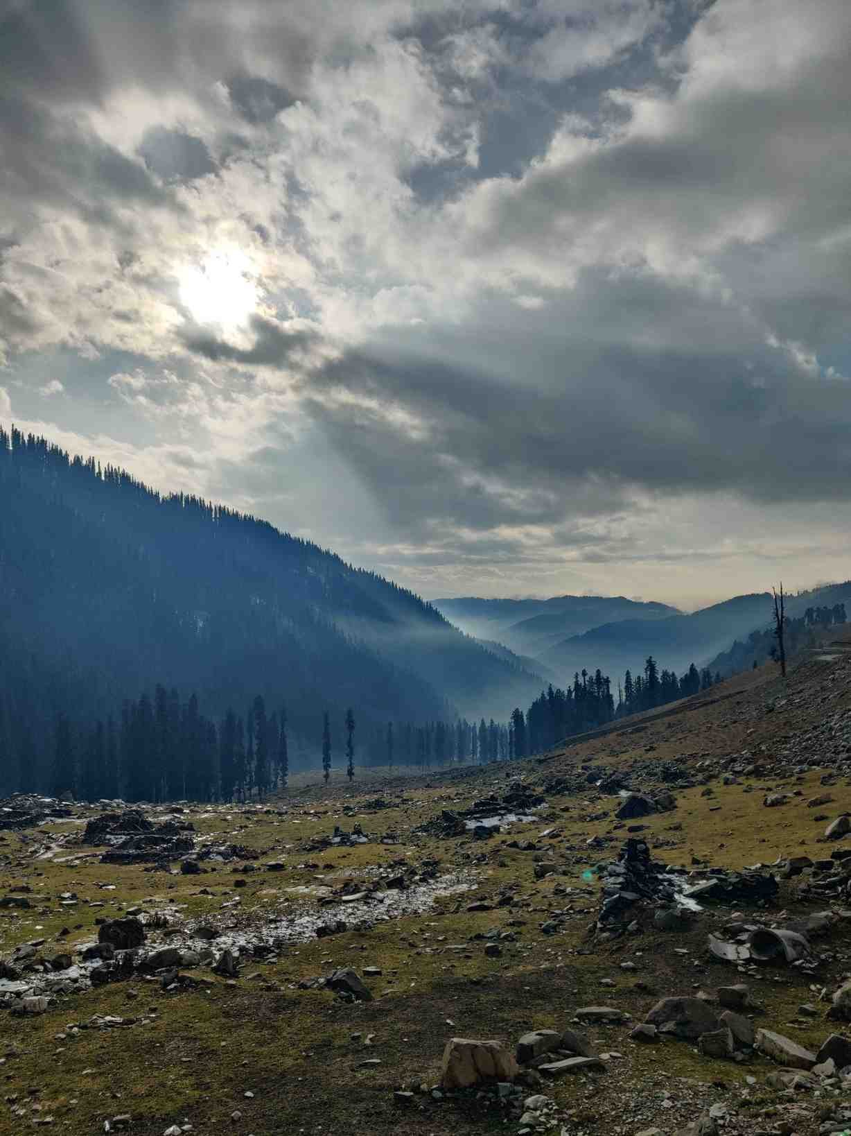 Sunlight breaking through dramatic clouds over a rocky Himalayan valley, illuminating pine-covered slopes and distant mountain ridges. 